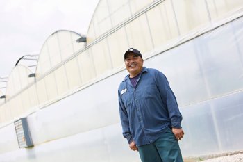 Migrant worker smiles at camera, standing in front of greenhouse